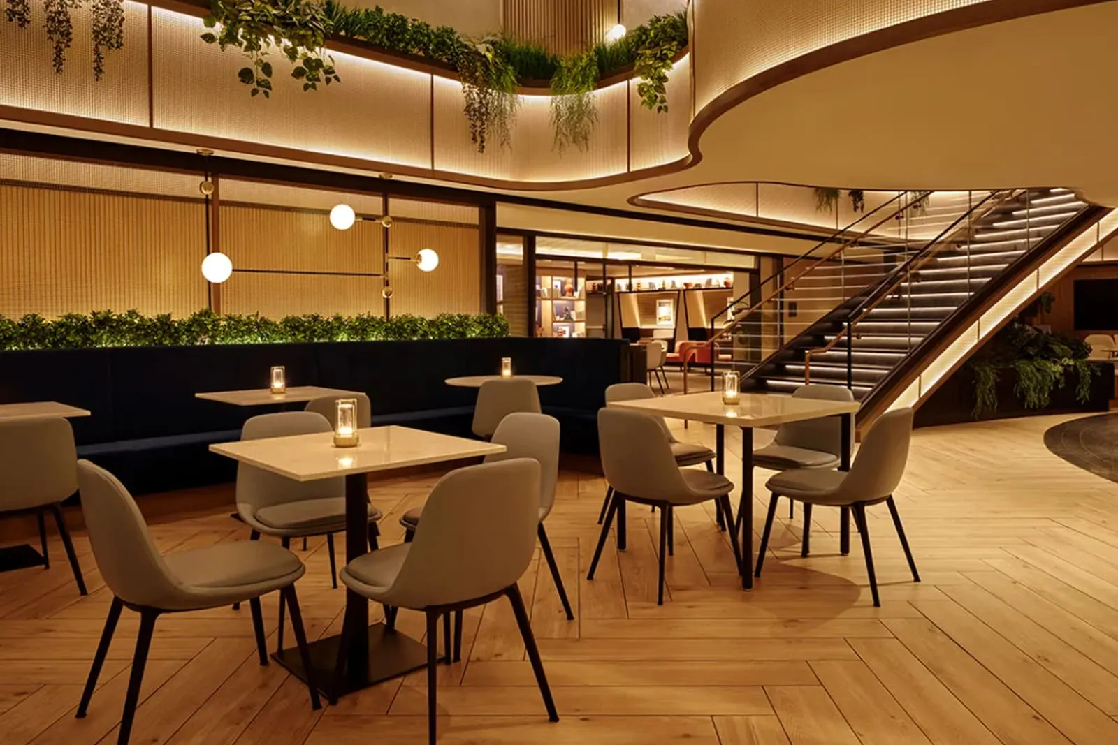 Dining tables with grey chairs and navy banquette under a plant-lined mezzanine staircase in the Chase Sapphire Lounge at LGA.