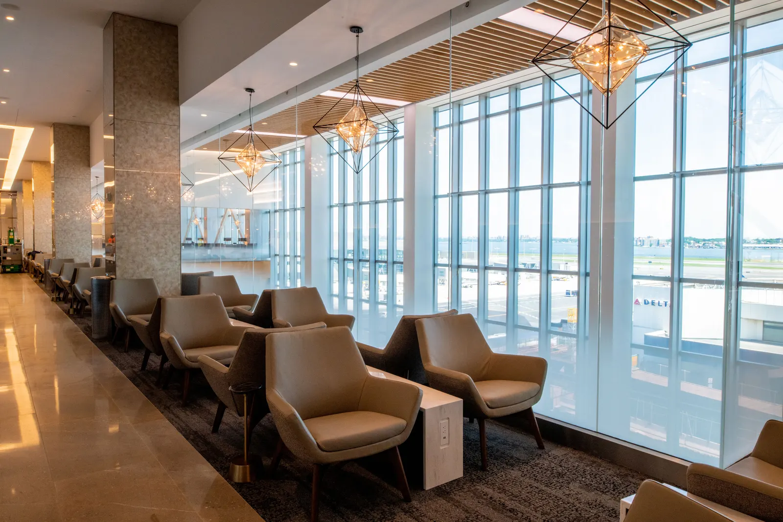 Tan armchairs with wood side tables under geometric pendants beside airfield windows in the Delta Sky Club at LaGuardia (LGA).