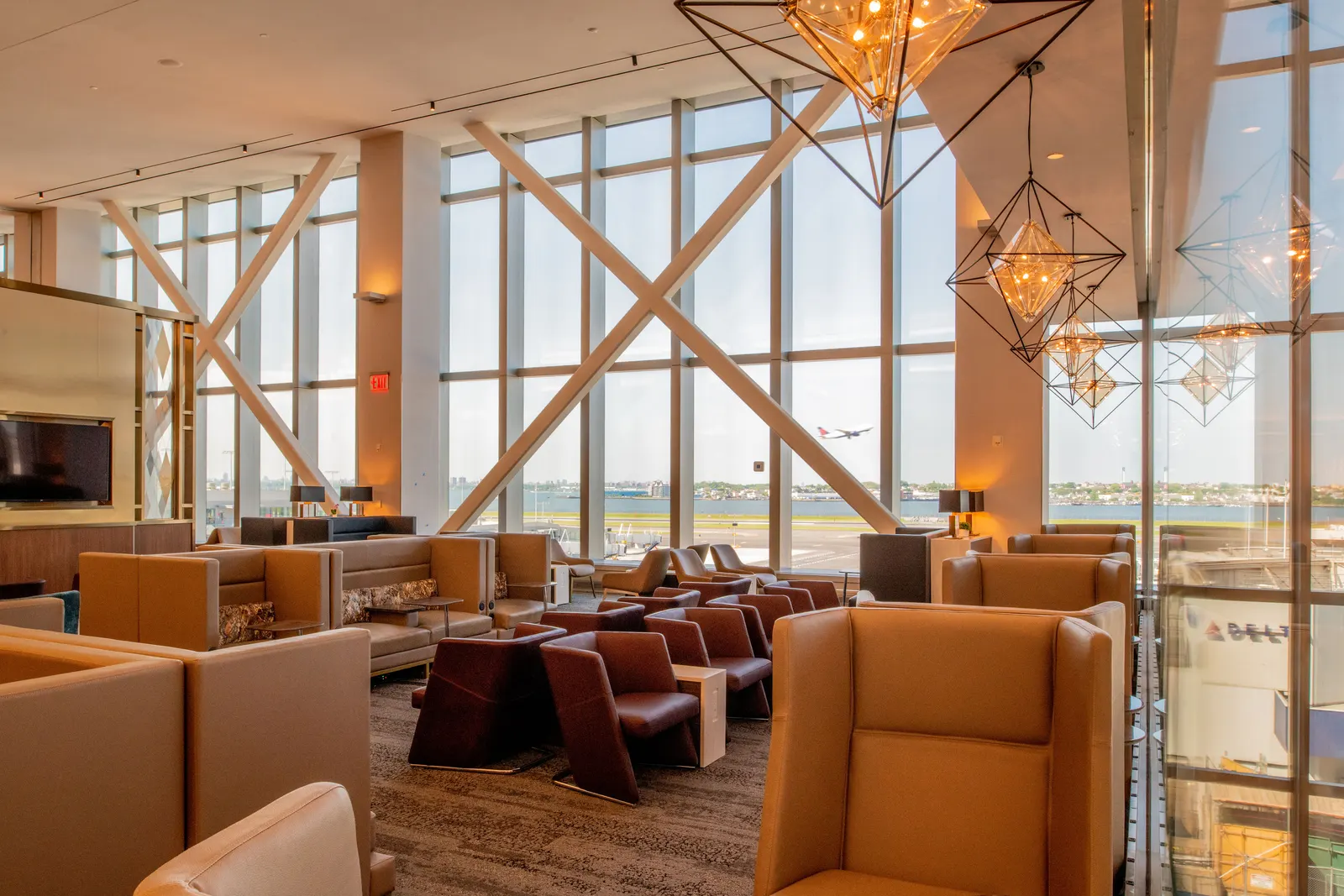 Beige privacy chairs and leather seating under geometric pendants with airfield windows in the Delta Sky Club at LaGuardia (LGA).