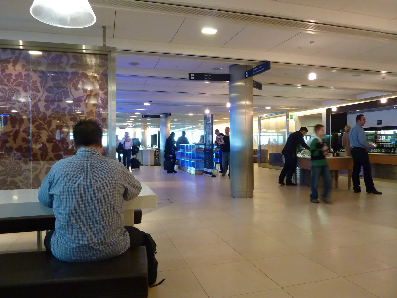 Open Galleries Club seating with purple damask glass partition, white tables, and a busy buffet island under pendant lights inside the British Airways Galleries Club Lounge at Heathrow Terminal 5 (LHR).