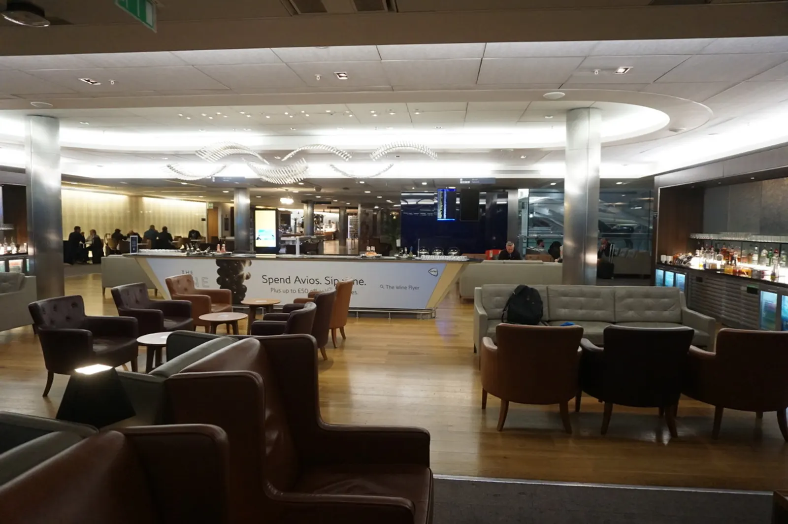 Leather seating, central refreshment counter, and Avios Wine Flyer banner inside the British Airways Galleries Club Lounge at Heathrow Terminal 5 (LHR).