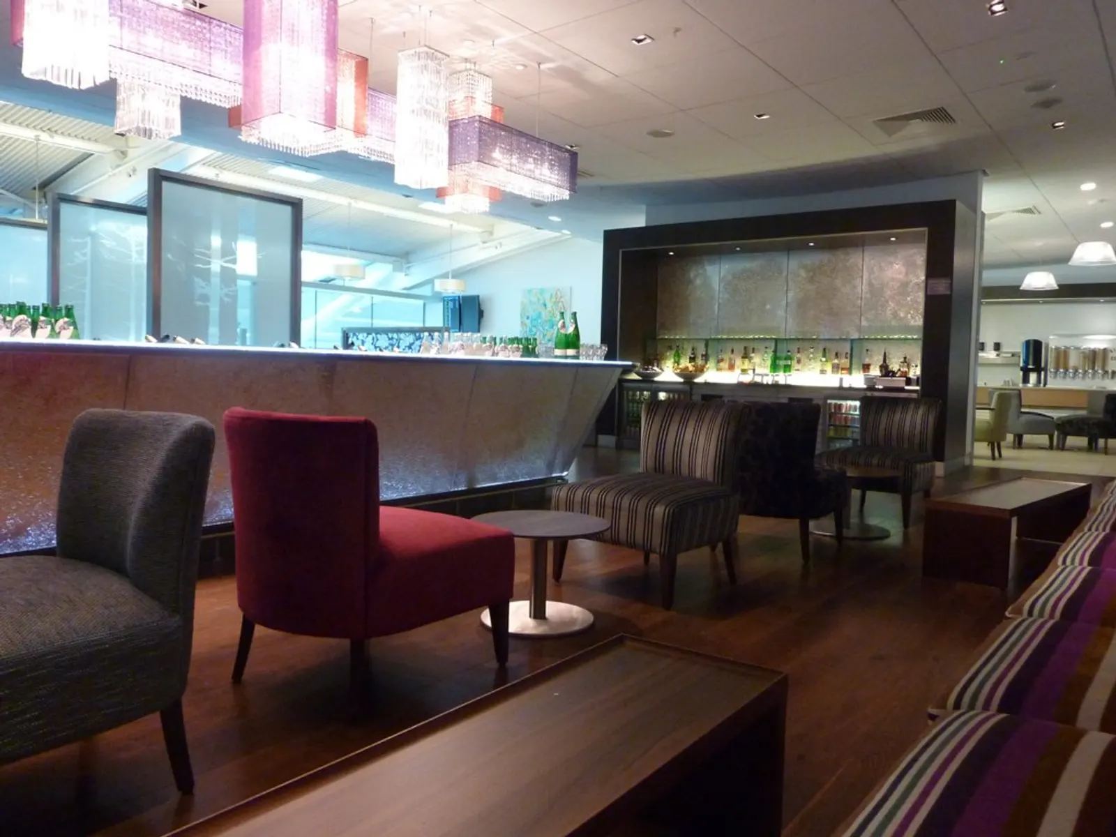 Bar with backlit bottle niche, striped sofas, and clustered pendant lights inside the British Airways Galleries Club Lounge South (T5 B satellite) at Heathrow Airport (LHR).