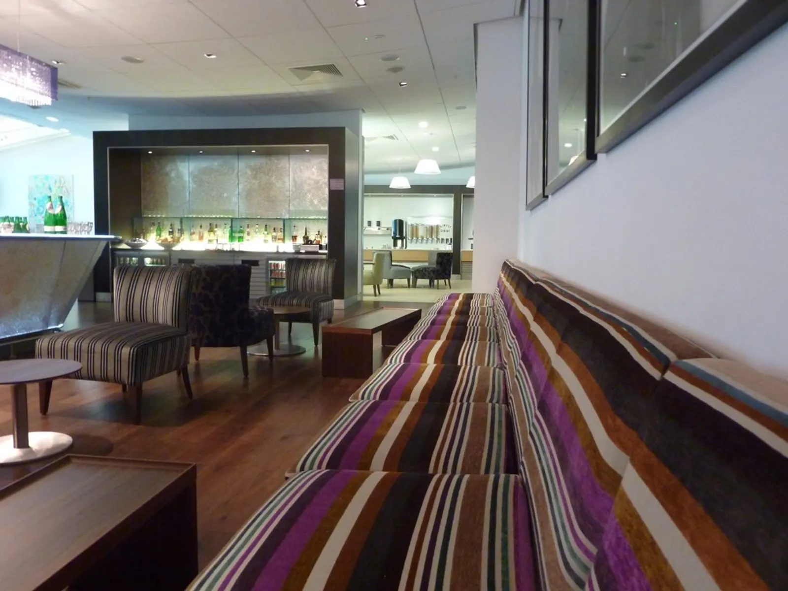 Purple-striped bench seating with dark wood tables facing a recessed liquor bar inside the British Airways Galleries Club Lounge South (T5 B satellite) at Heathrow Airport (LHR).