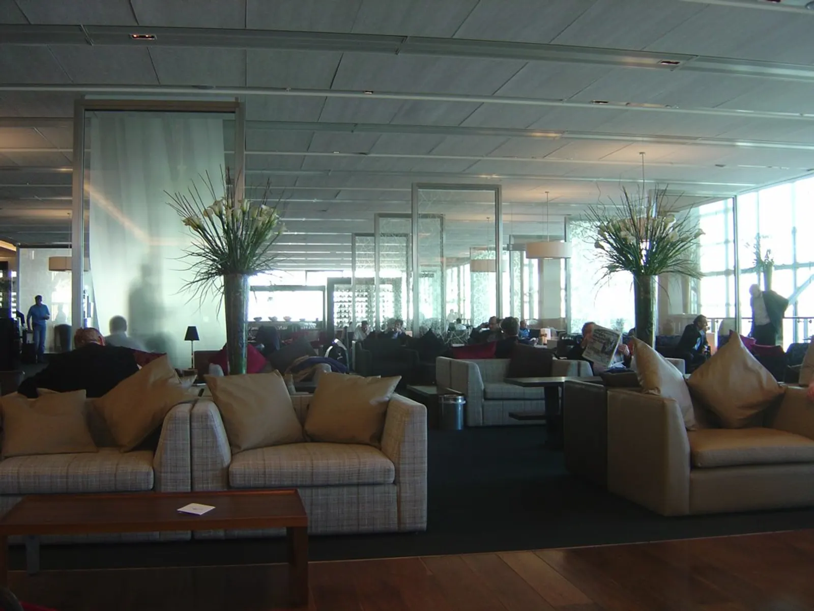 Beige armchair seating with tall lily arrangements, glass partitions, and window light inside the British Airways Galleries First Lounge at Heathrow Terminal 5 (LHR).