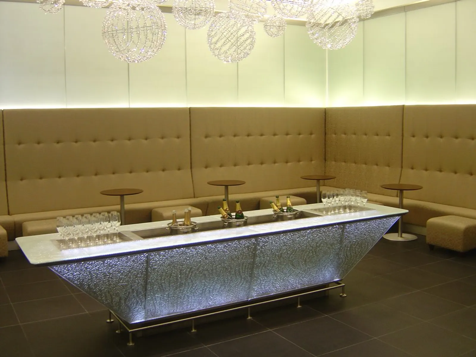 Illuminated champagne bar with ice buckets and flute trays beside tufted banquettes inside the British Airways Galleries First Lounge at Heathrow Terminal 5 (LHR).