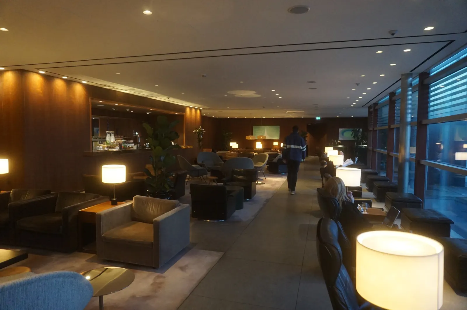 Warm lamplit seating aisle with dark wood bar niche and floor-to-ceiling windows inside the Cathay Pacific First and Business Class Lounge at Heathrow Airport (LHR).