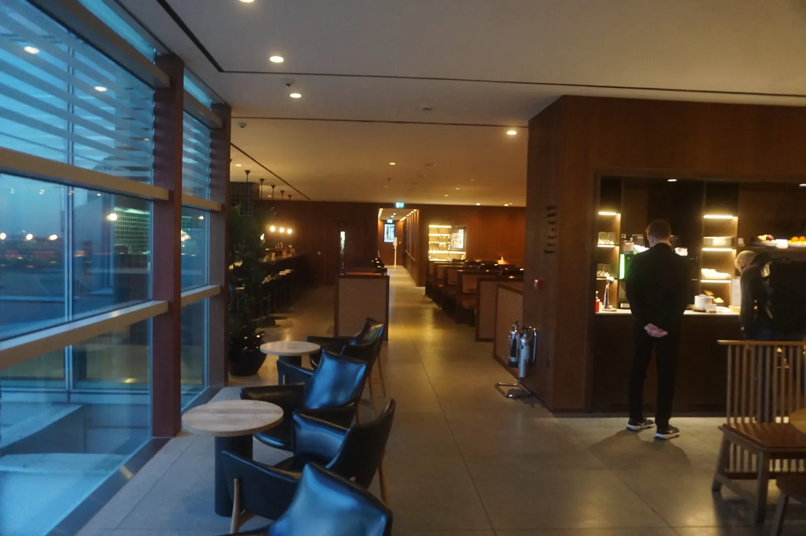 Black leather window seating opposite a dark-wood service counter and booth partitions inside the Cathay Pacific First and Business Class Lounge at Heathrow Airport (LHR).