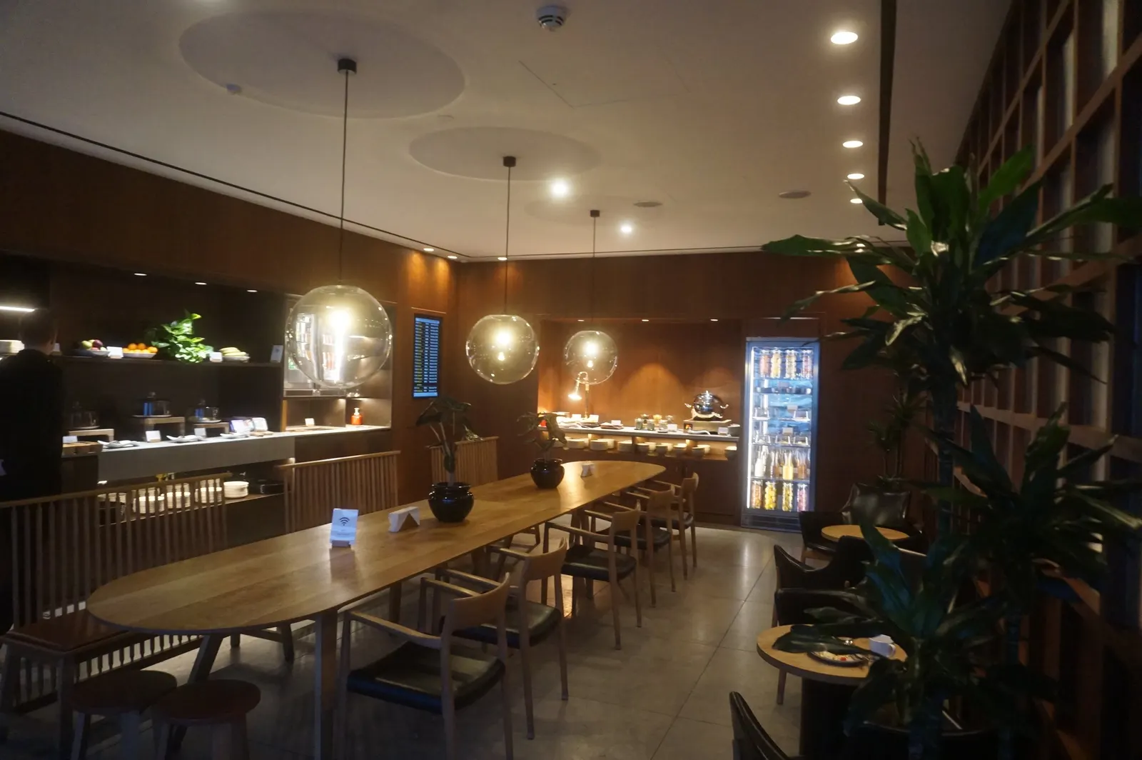 Long wooden communal dining table under spherical glass pendants beside a wood lattice divider and beverage fridge inside the Cathay Pacific First and Business Class Lounge at Heathrow Airport (LHR).
