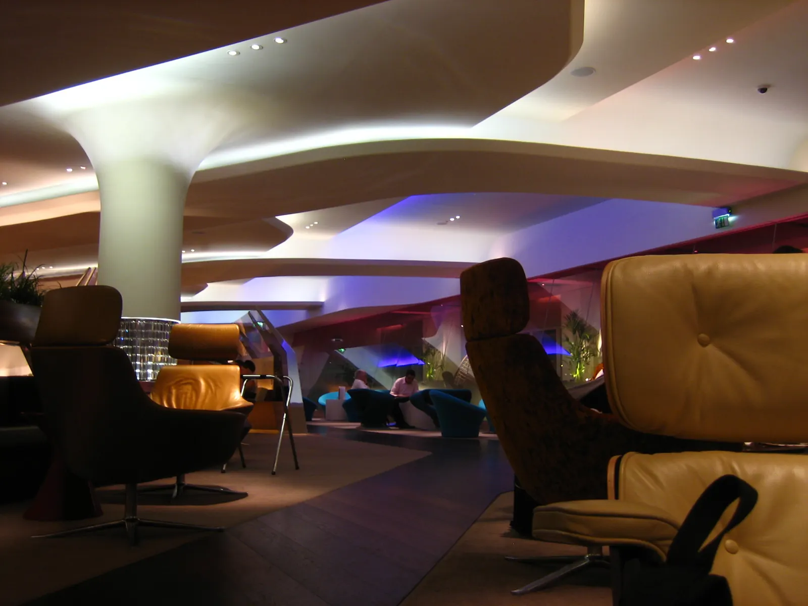 Curved white layered ceiling with purple-blue accent lighting above tan and blue lounge chairs and glass partitions inside the Virgin Atlantic Clubhouse at London Heathrow Airport (LHR) Terminal 3.