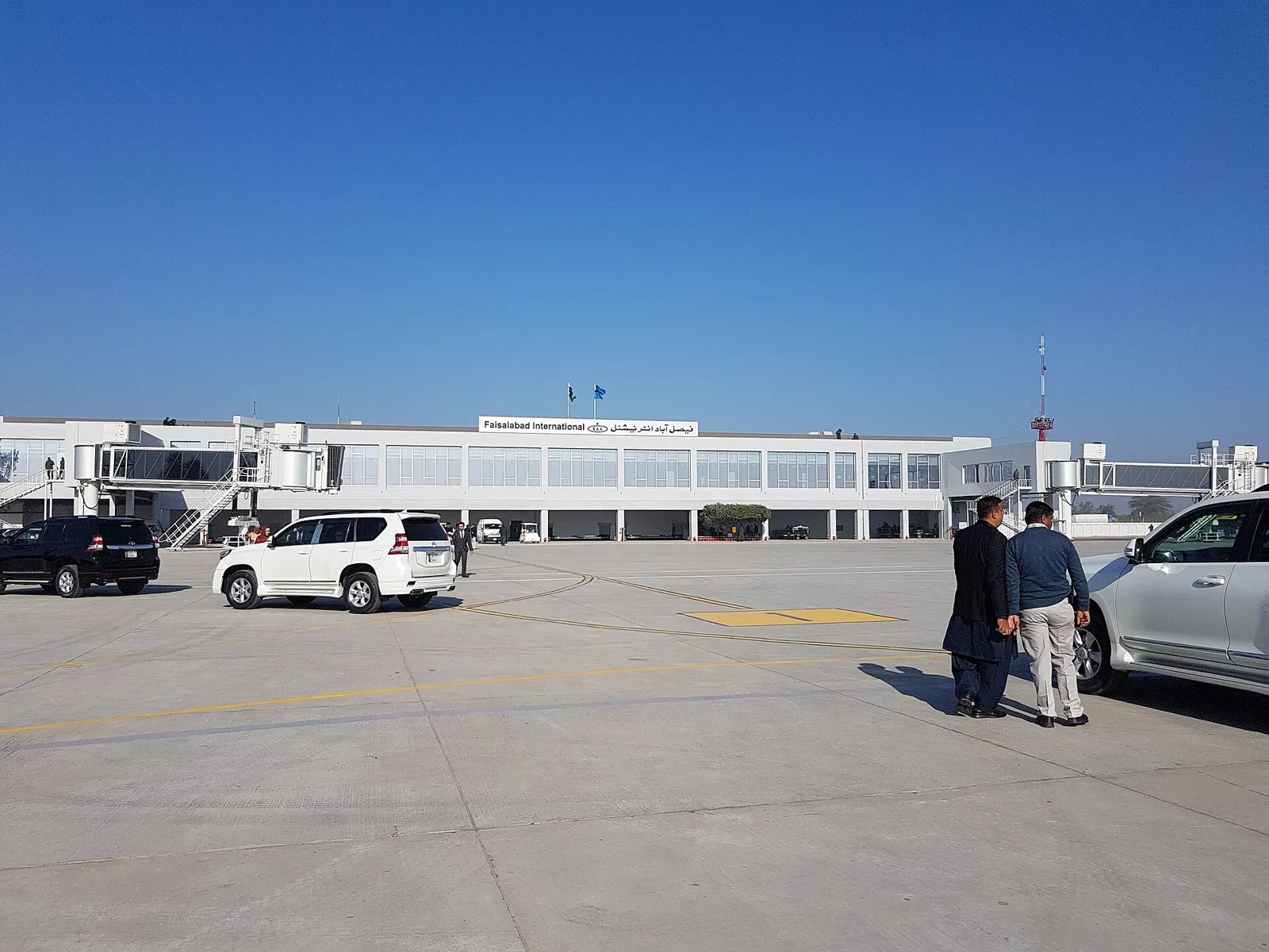 Long white single-storey terminal building with a control tower behind, seen across a wide paved forecourt with parked cars under a clear blue sky