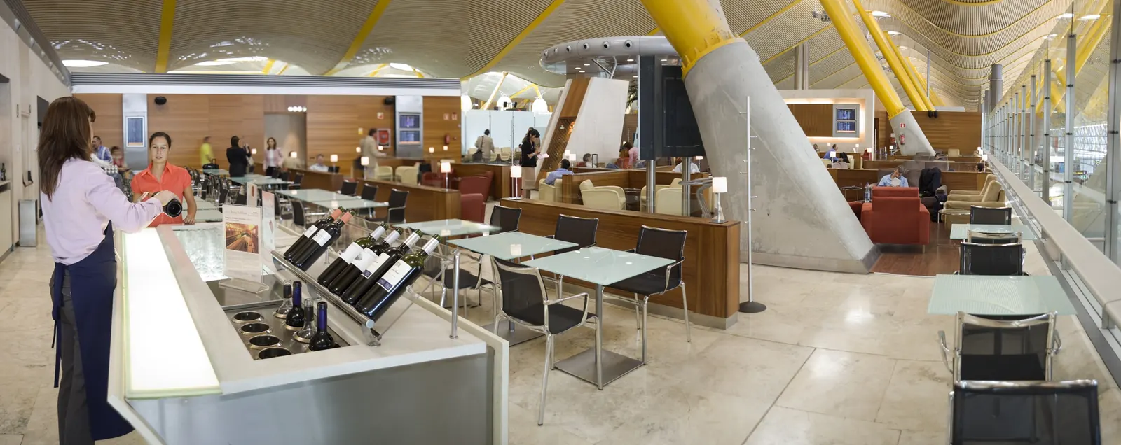 Wine bar counter with staff pouring, glass dining tables, and red and beige lounge seating under the T4 wave ceiling inside Iberia Premium Lounge Dalí at Madrid-Barajas (MAD).