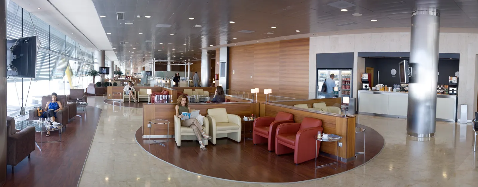 Wide lounge seating with cream and red armchairs, circular wood floor inlays, and a refreshment counter beside tall windows inside Iberia Premium Lounge Velázquez at Madrid-Barajas Terminal 4 (MAD).
