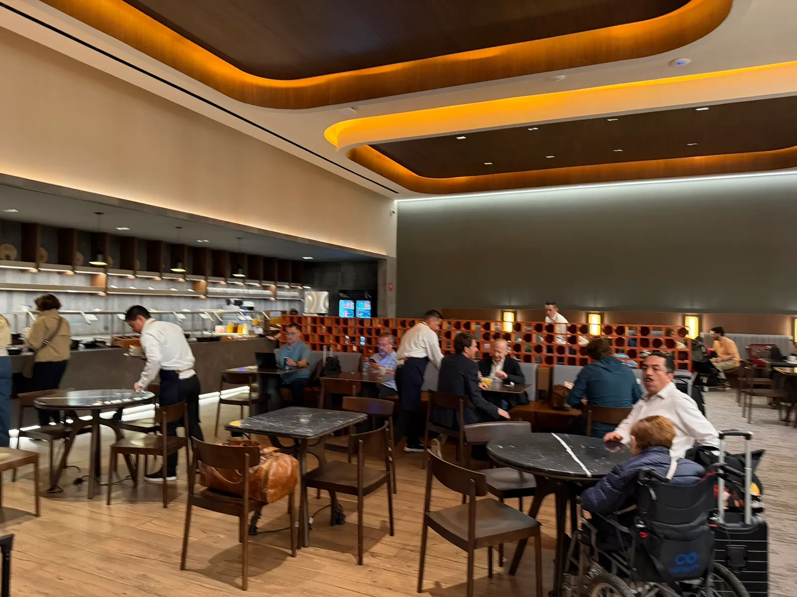 Dining tables with wood chairs beside a buffet counter and orange lattice screen under warm recessed ceiling lights inside Aeroméxico Salón Premier Nacional at Mexico City International (MEX).
