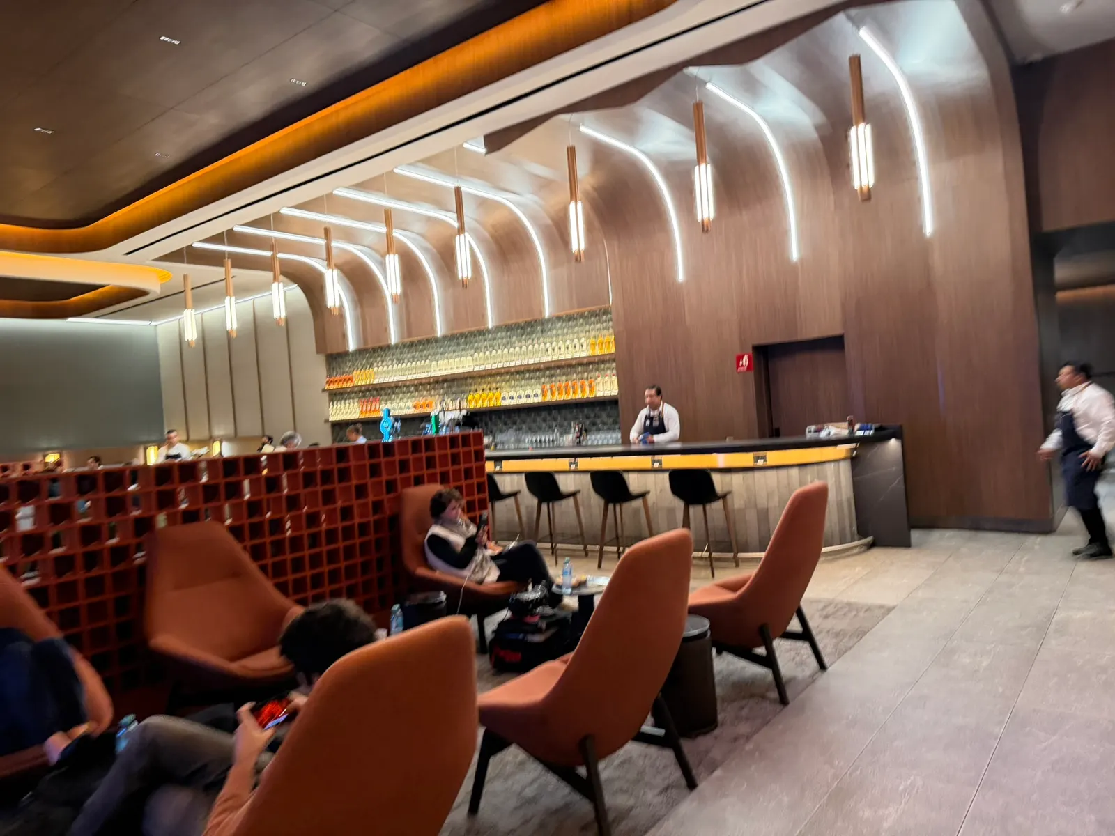 Terracotta lounge chairs facing a curved wood-ribbed bar with pendant lights and a red lattice partition inside Aeroméxico Salón Premier Nacional at Mexico City International (MEX).