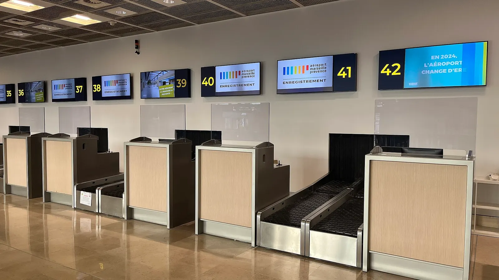 Row of 2024 Terminal 1 check-in desks numbered 35 to 42 with Aéroport Marseille Provence screens and wooden bag belts.