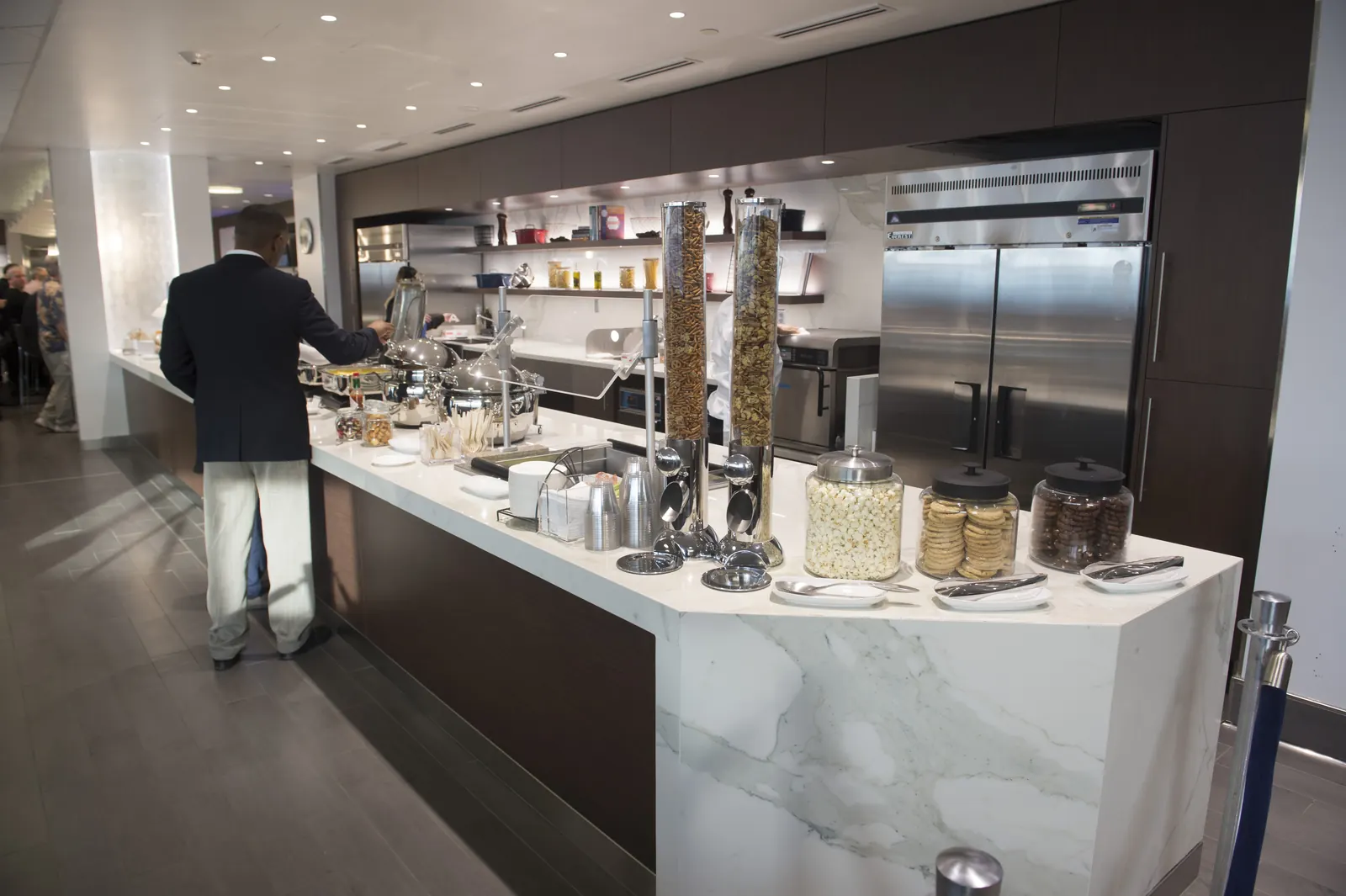 Marble buffet counter with cereal dispensers, snack jars, and stainless warmers under dark wood cabinetry inside the Delta Sky Club at Minneapolis-Saint Paul International Airport (MSP).