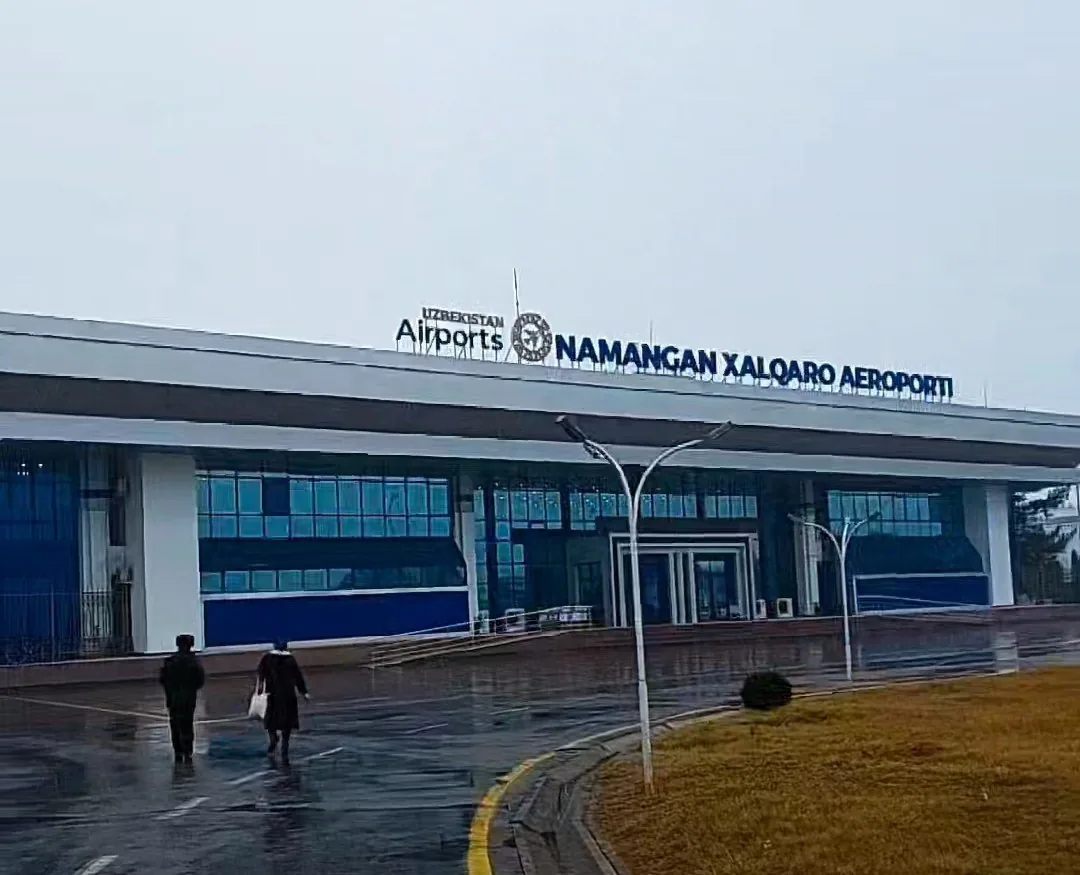 Landside front of the passenger terminal at Namangan International Airport with blue glass facade and the 'Uzbekistan Airports – Namangan xalqaro aeroporti' sign on a rainy day