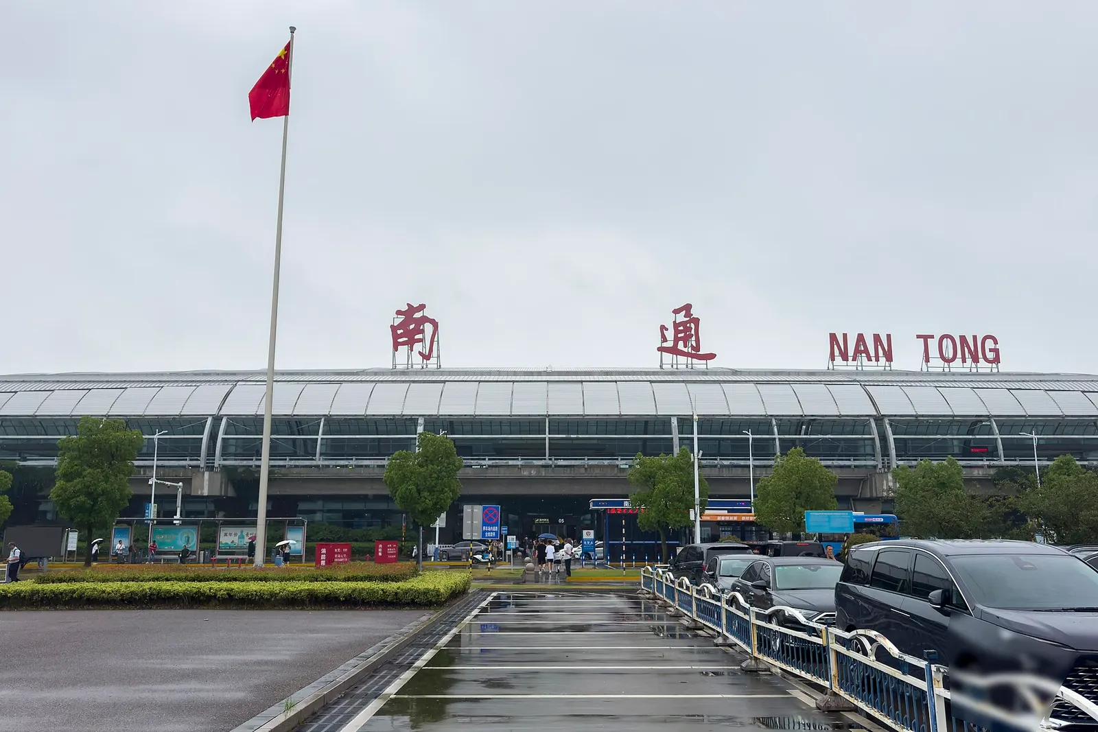 Wet curb and glass façade of Nantong Xingdong Terminal 3 with 南通 NAN TONG letters and a Chinese flag on a tall pole, 19 June 2026.