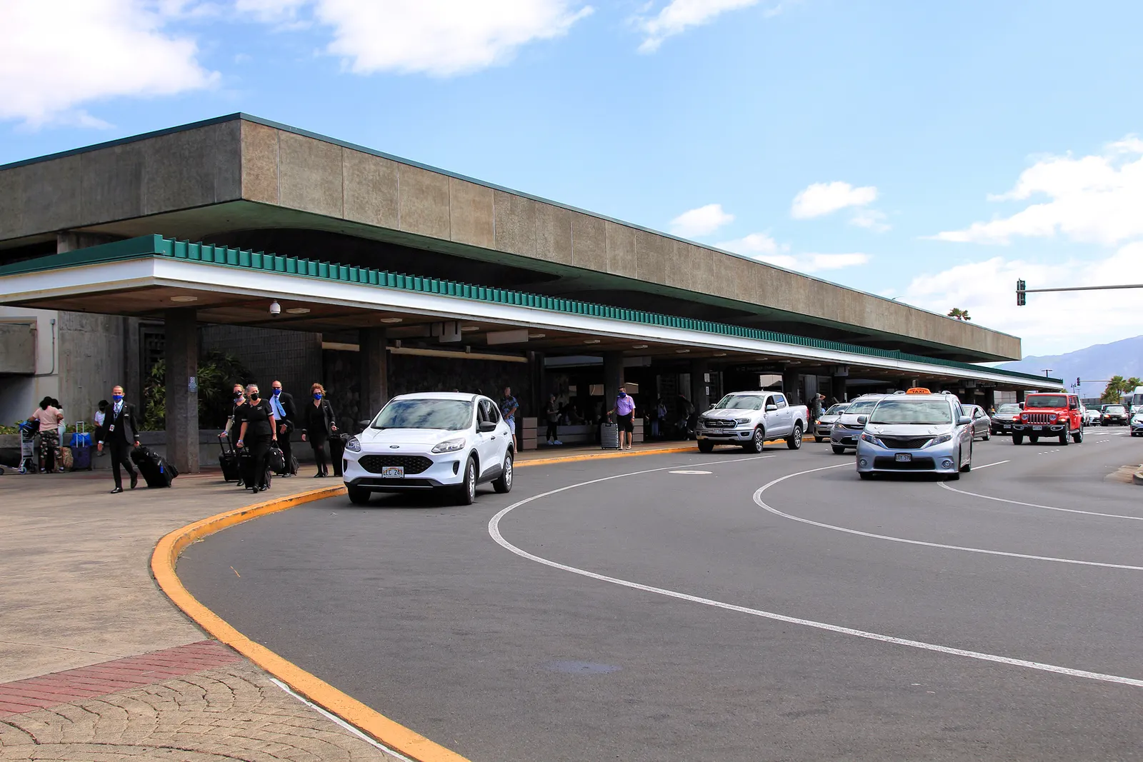Landside curb and concrete canopy at Kahului Airport in 2021, with Hawaii plates and masked travelers on the sidewalk.