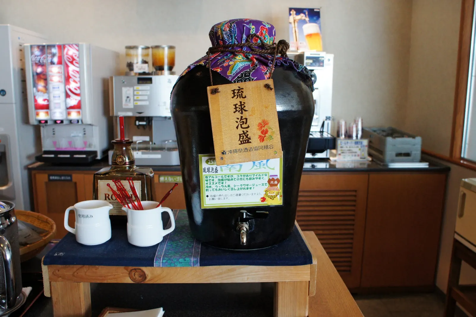 Ceramic drink jar with Japanese sign beside Coca-Cola and coffee machines on a wood counter inside the JAL Sakura Lounge at Naha Airport (OKA).