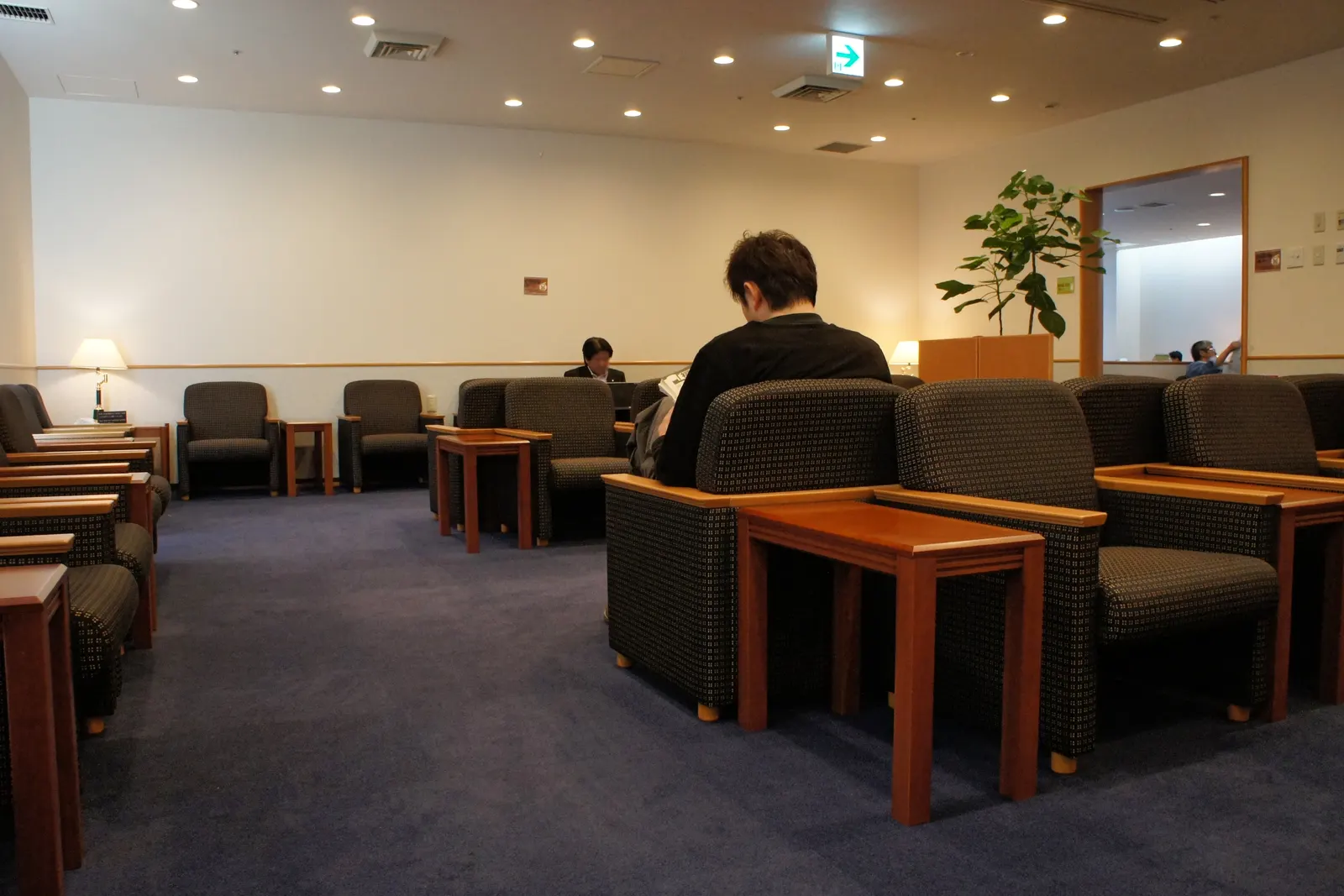 Rows of charcoal armchairs with reddish wood side tables on navy carpet under recessed ceiling lights inside the JAL Sakura Lounge at Naha Airport (OKA).