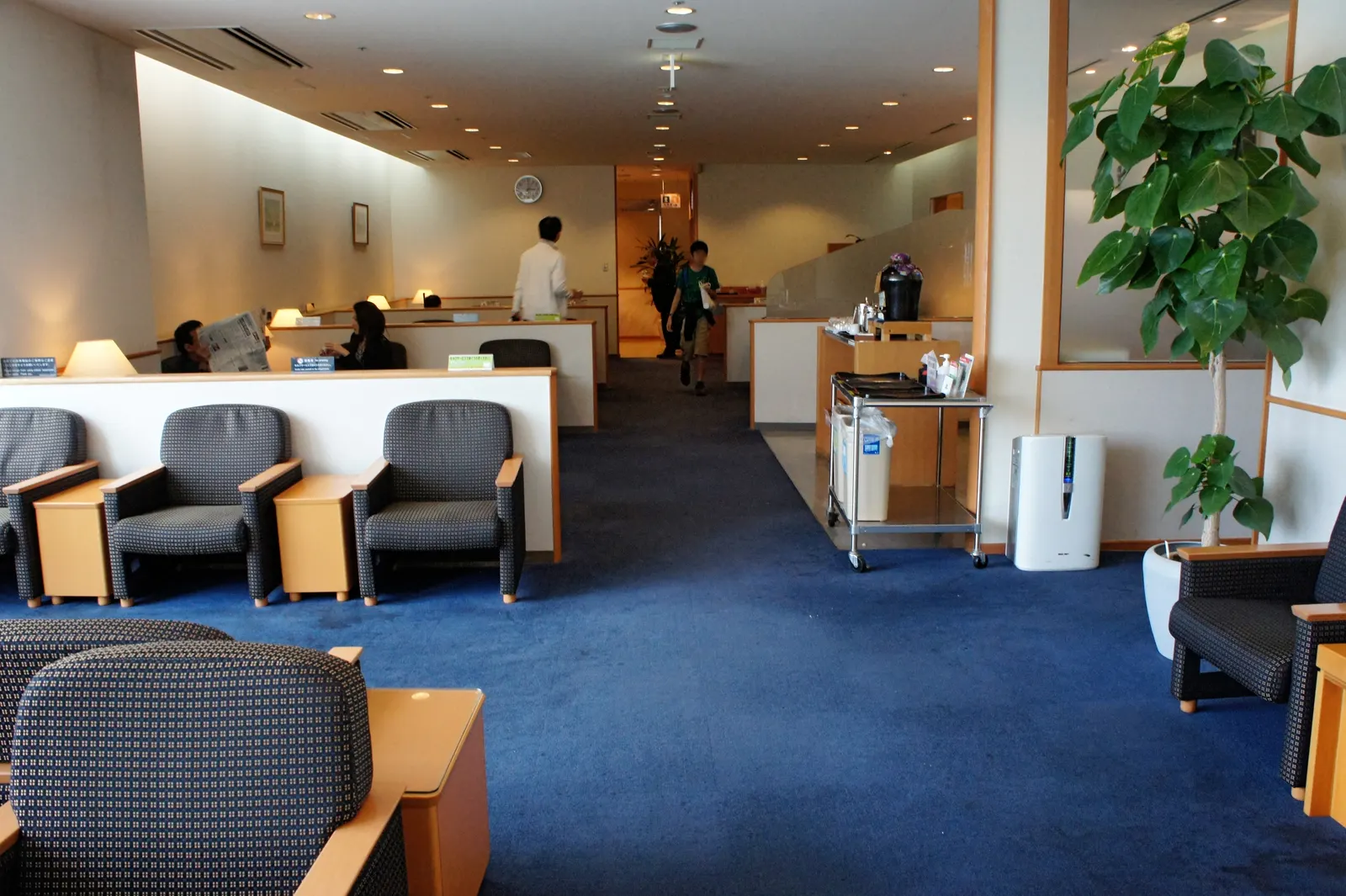 Central aisle with partitioned seating clusters, a wood refreshment counter, and soft ceiling lighting inside the JAL Sakura Lounge at Naha Airport (OKA).
