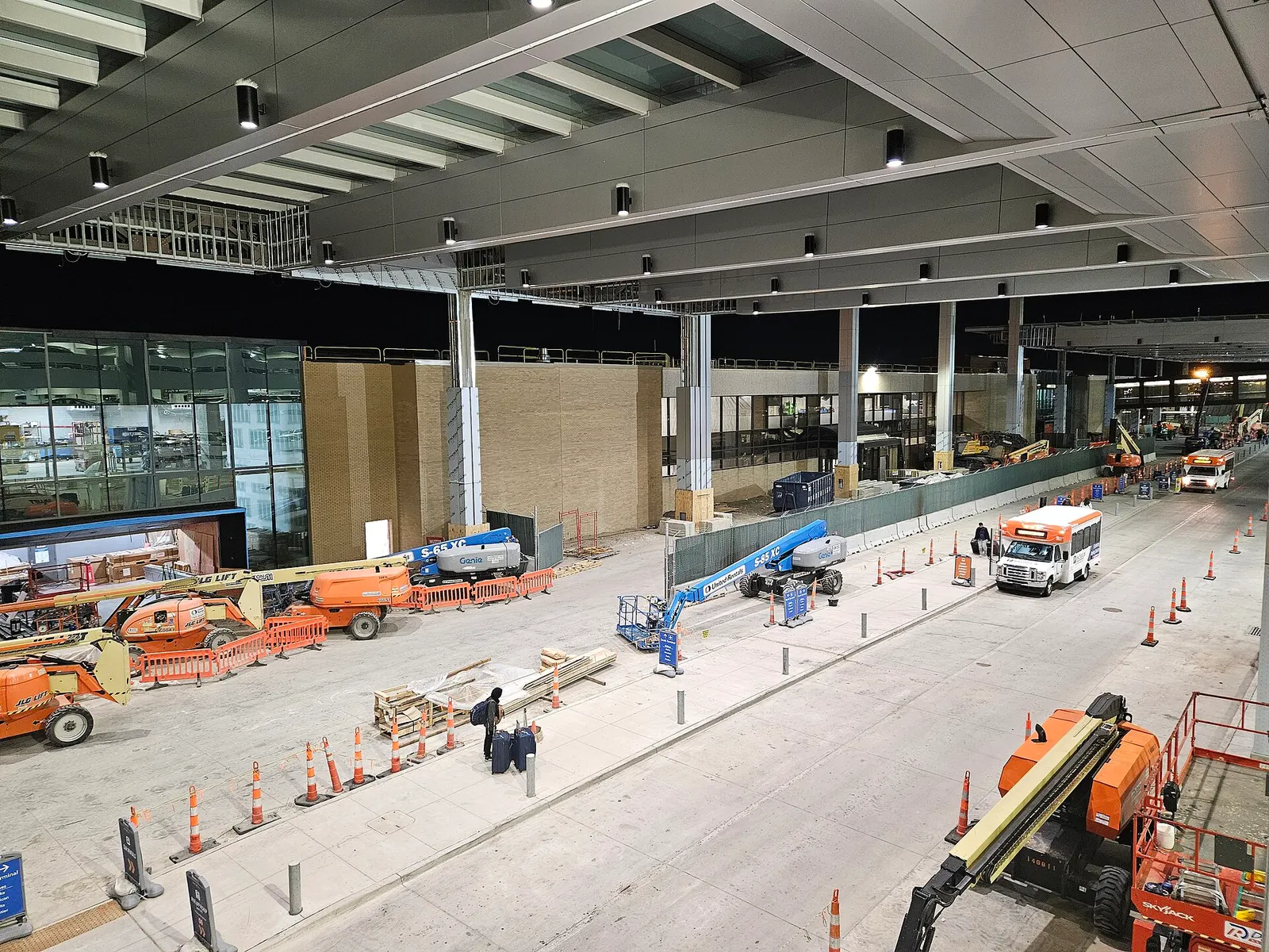 Night view of Build OMA construction at the Eppley terminal curb, with boom lifts, a green construction fence, an overhead canopy, shuttle buses, and a traveler with rolling bags.