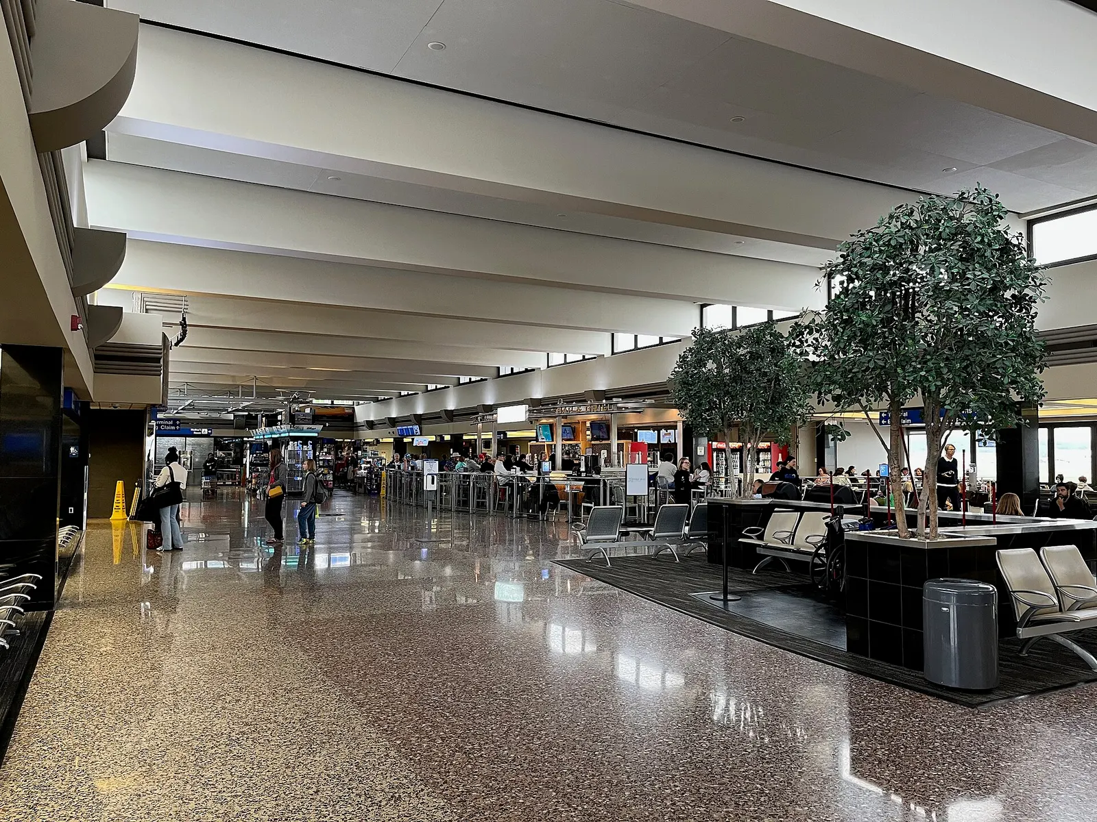 2025 airside concourse at Eppley Airfield with polished terrazzo, a central bar and newsstand, tree planters, and a blue sign pointing to B Gates and baggage claim.