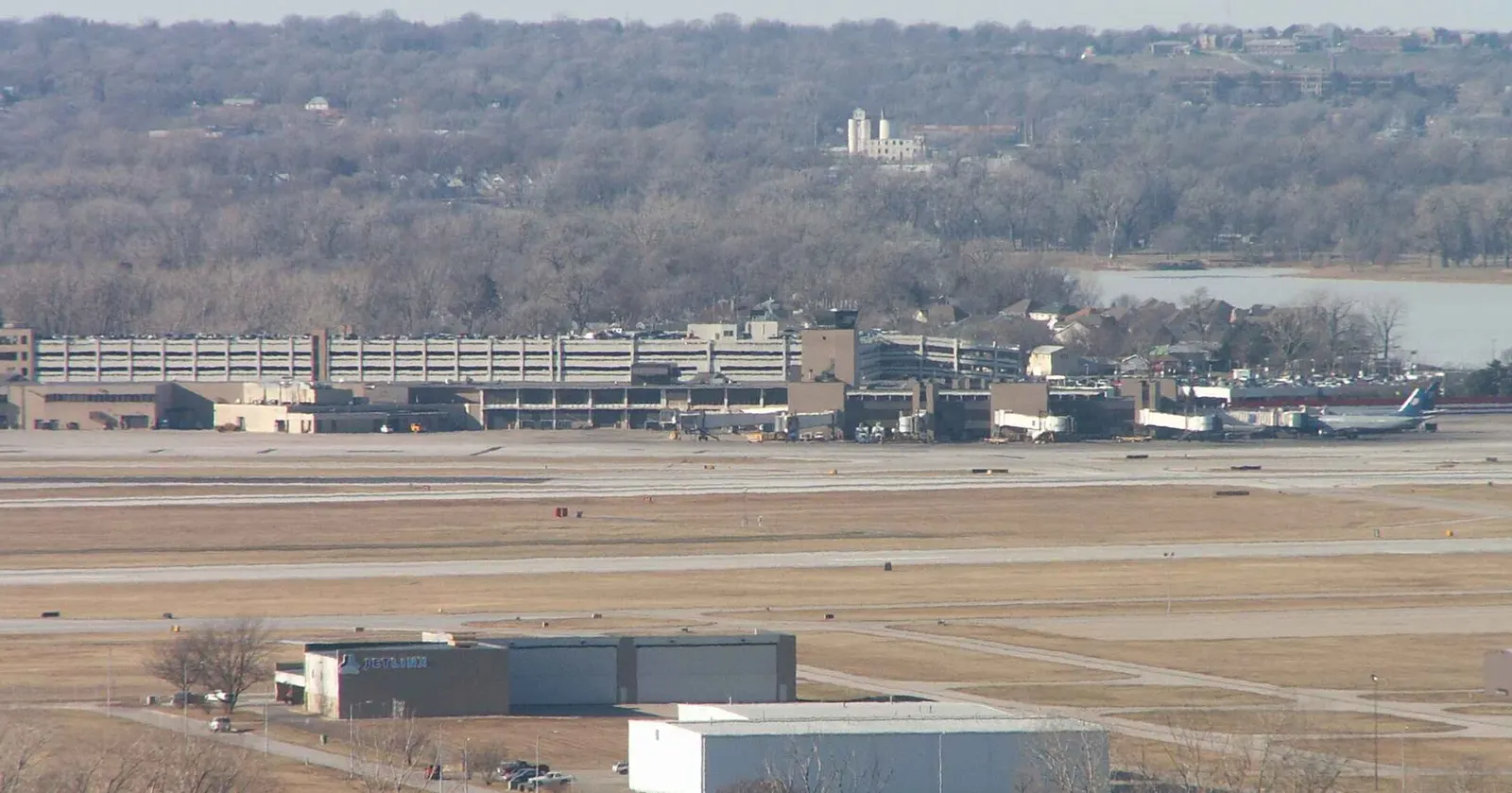 Elevated view of the Eppley Airfield terminal, parking garage, and jet bridges with Carter Lake behind the field and a Jet Linx hangar in the foreground.