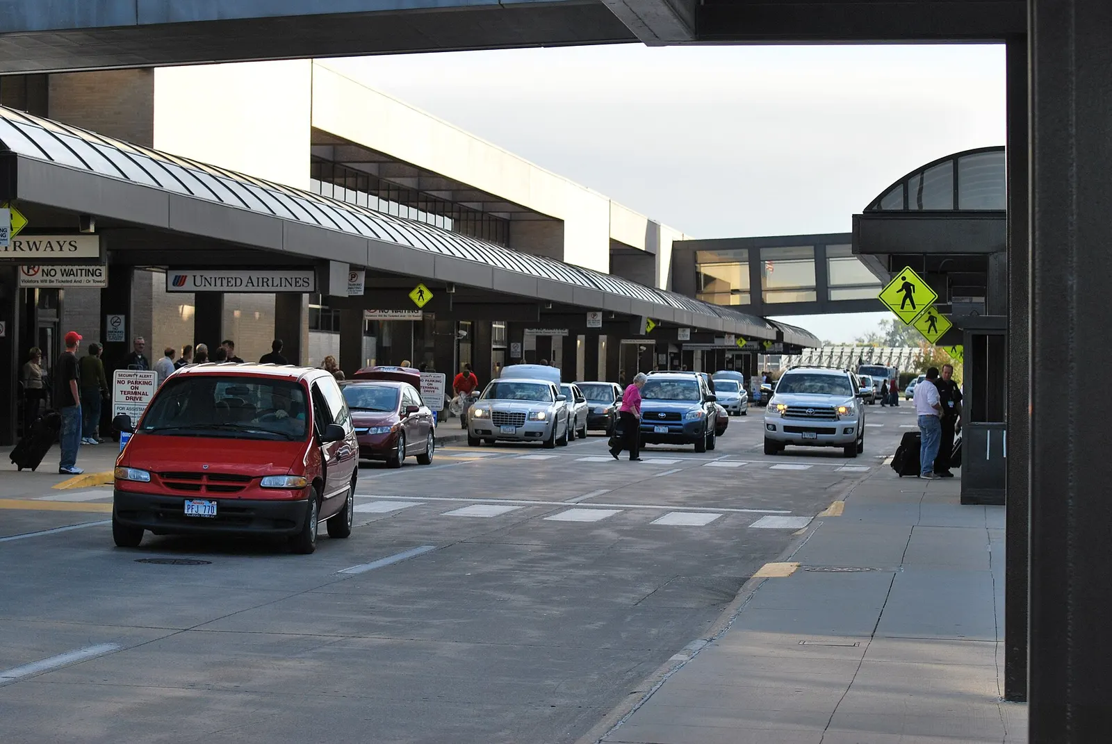 Daylight curb at Eppley Airfield showing United and US Airways canopy signs, a skywalk to the garage, no-waiting posts, and cars at the crosswalk.