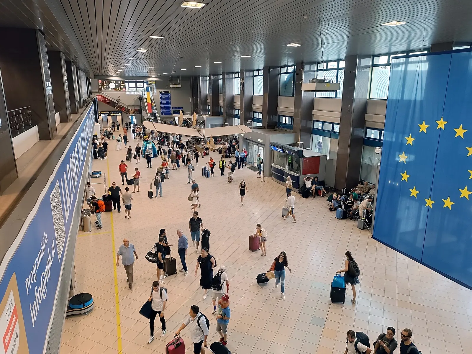 Busy check-in hall at Henri Coandă International Airport with a vintage aircraft hanging above the floor and a large EU flag on the wall.