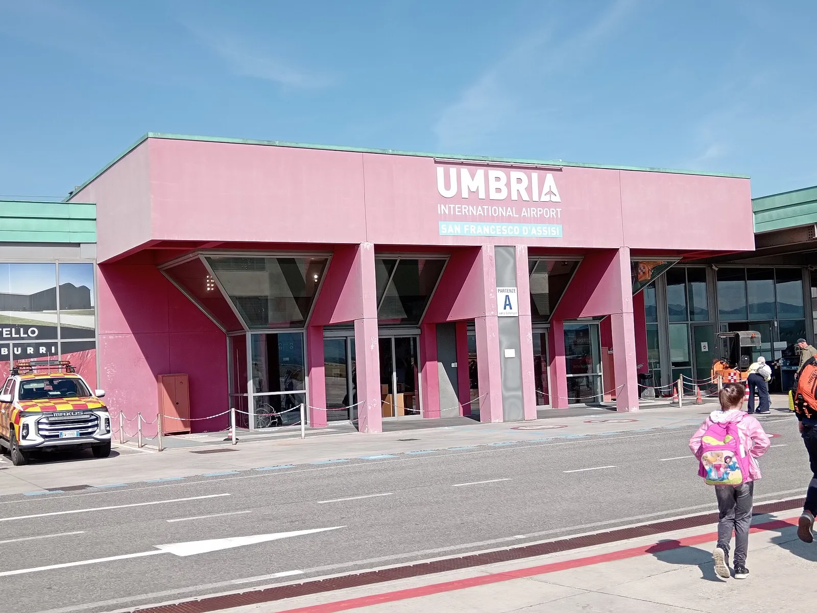 Red and pink pavilions of the Perugia airport terminal with the UMBRIA International Airport sign, seen from the landside road