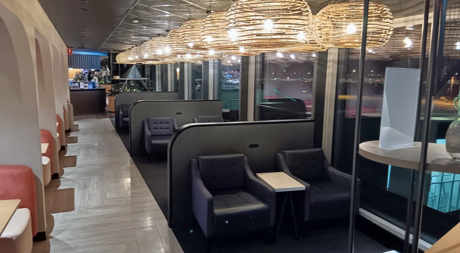 Semi-private blue armchair booths and peach banquettes beside floor-to-ceiling windows at night inside the International Aspire Lounge at Perth Airport (PER).
