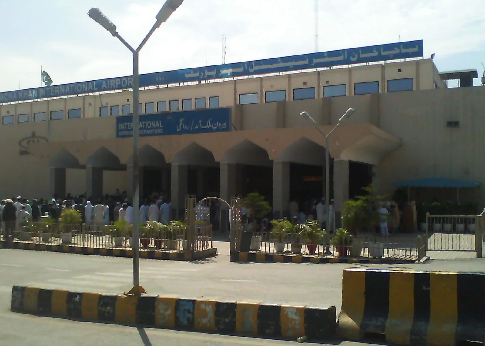Front of Bacha Khan International Airport in Peshawar: a beige terminal with a row of arches, a blue 'International Arrival & Departure' sign in English and Urdu, and the airport's name along the roof, with passengers waiting behind a railing and striped kerbstones in the foreground