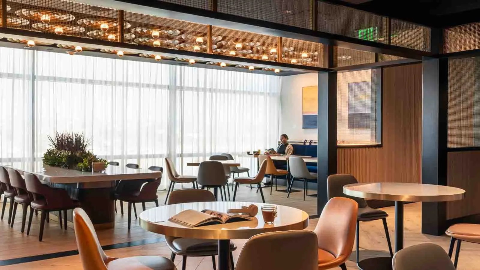 Dining tables and communal seating by curtained windows in the Chase Sapphire Lounge by The Club at San Diego International Airport (SAN).