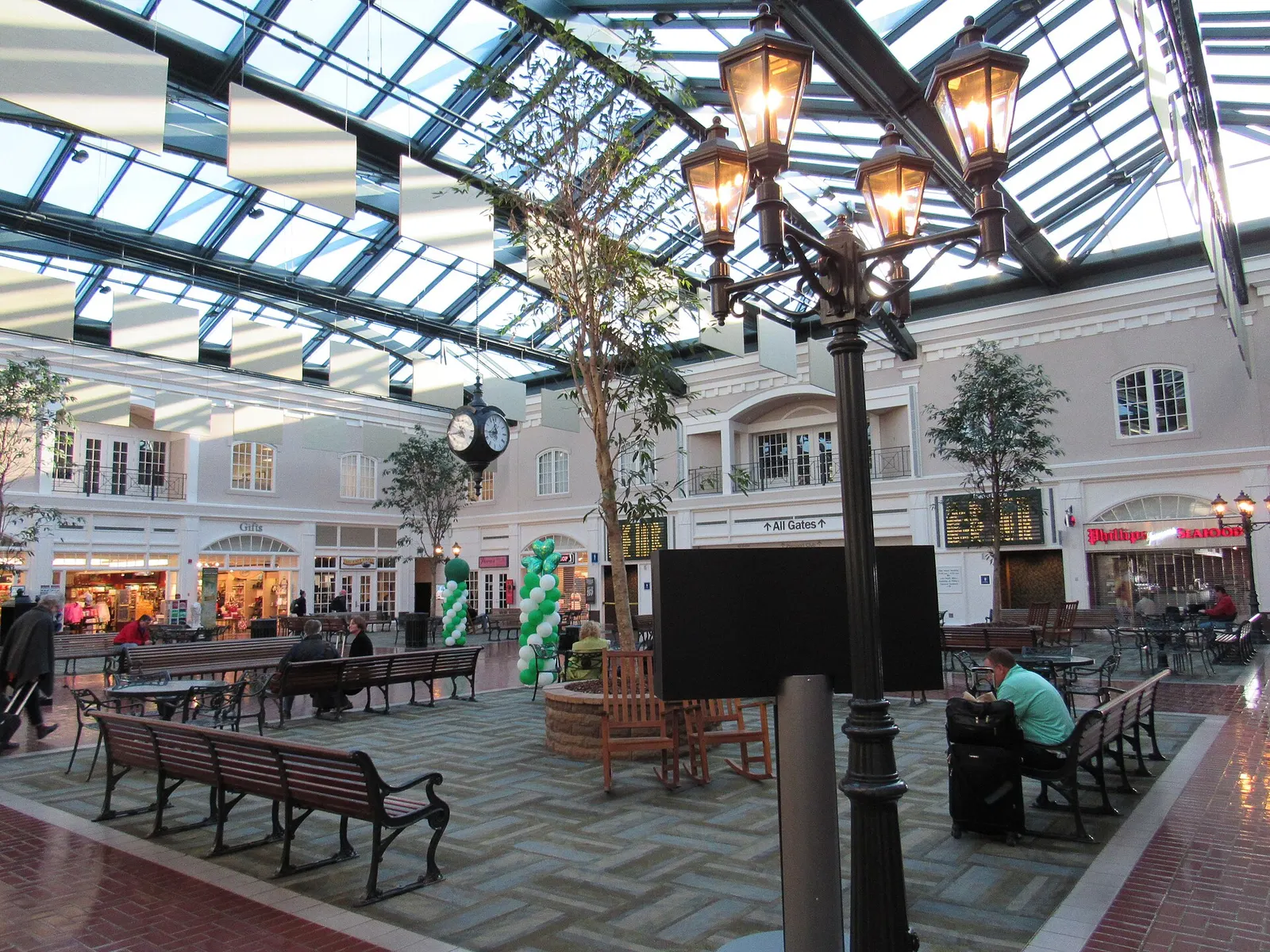 Savannah Square atrium at SAV: glass roof, hanging clock, park benches, Gifts shop, Phillips Seafood, and an All Gates sign toward the concourse.