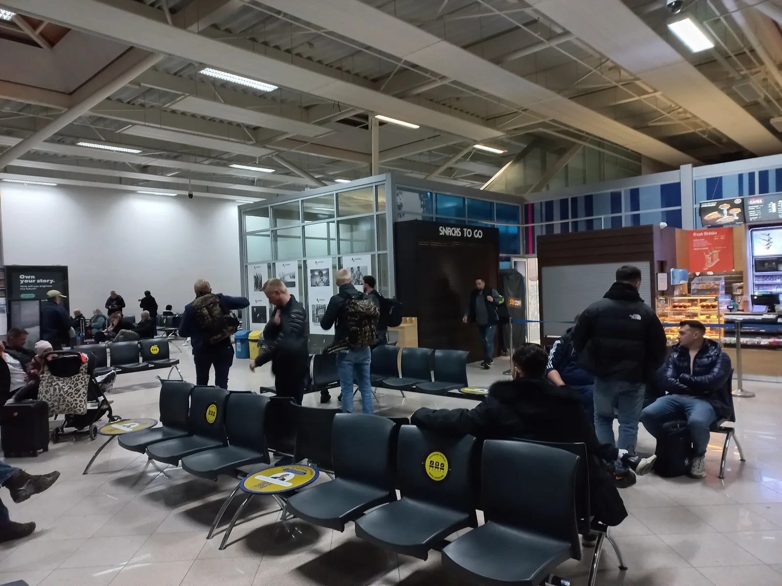 Passenger waiting area at Sibiu International Airport with rows of black seats, a 'Snacks to go' kiosk and a bakery and coffee counter