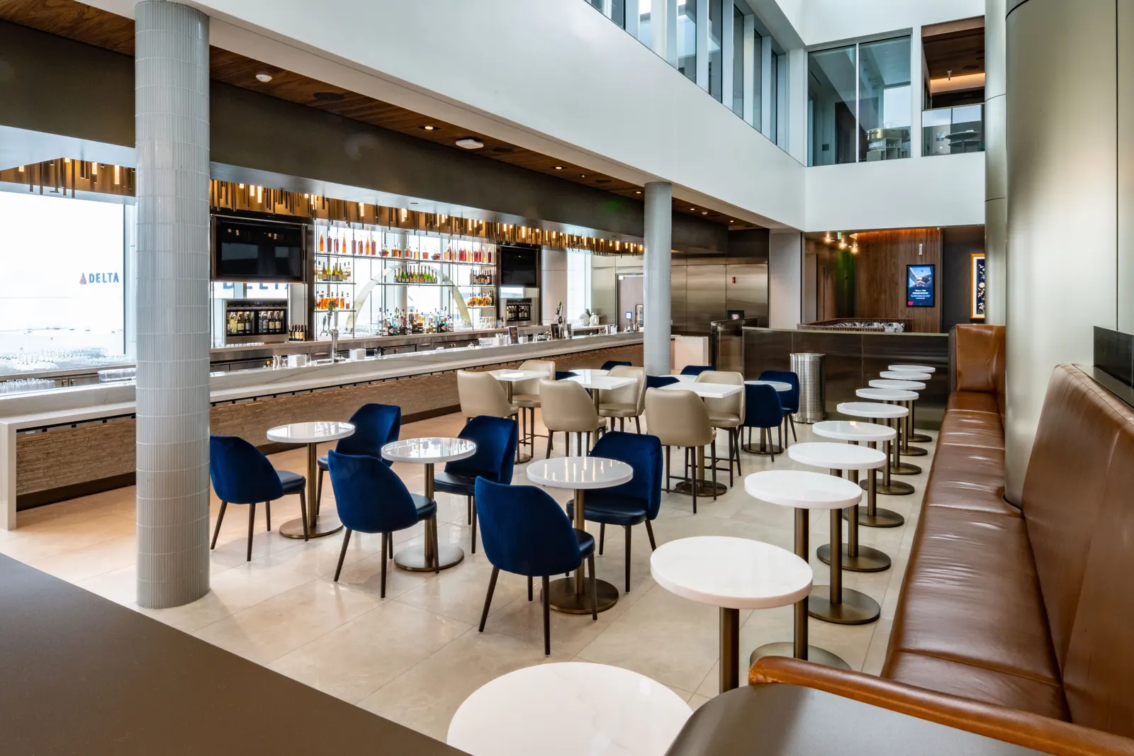 Blue and tan armchairs at marble tables beside a long bar with airfield windows in the Delta lounges complex at Seattle-Tacoma (SEA).