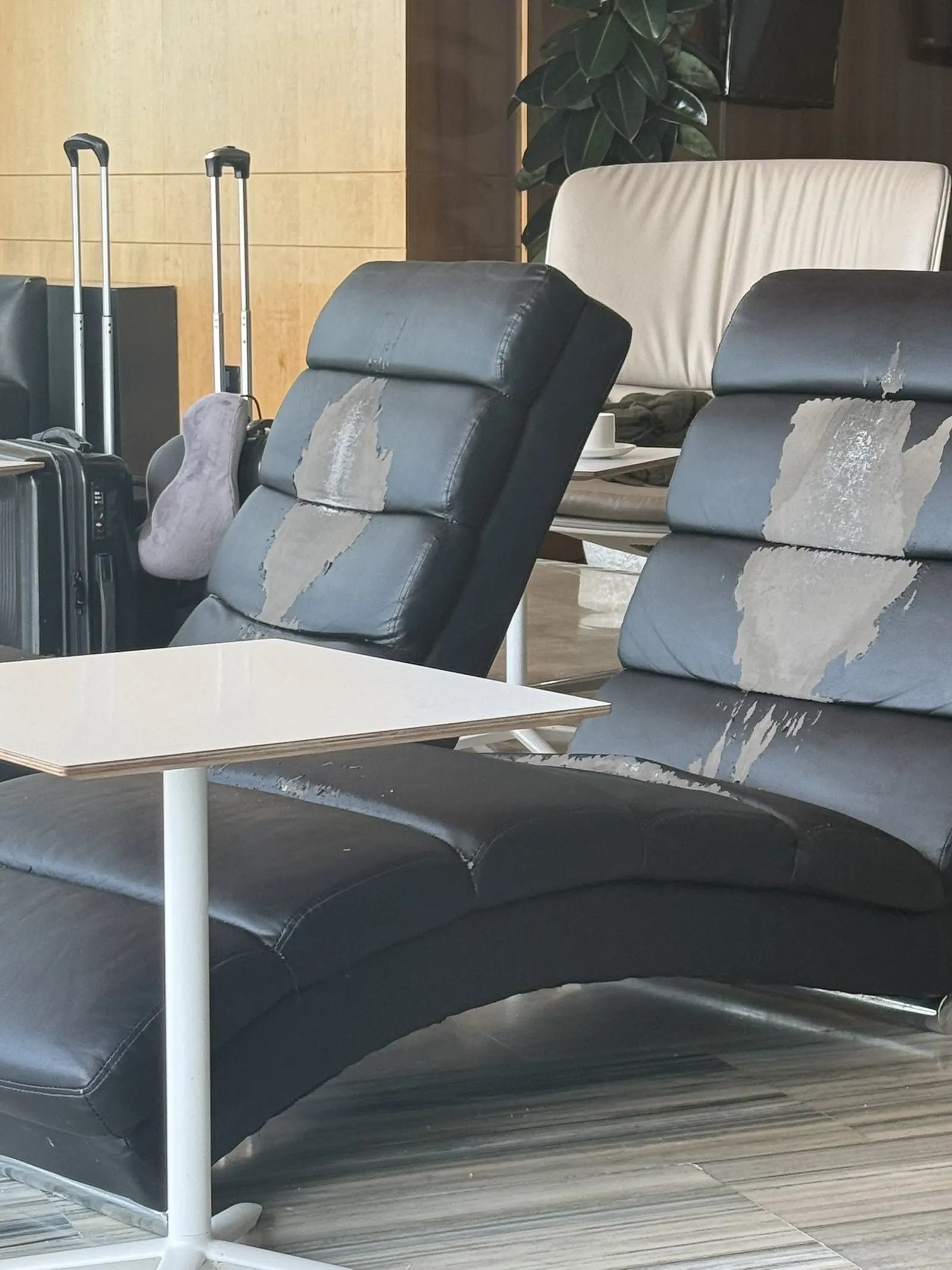 Two black contoured faux-leather recliner chairs with large patches of covering peeled from the headrests and backs, beside a white laminate side table on marble-look flooring in Montaña Roja VIP Lounge at TFS.