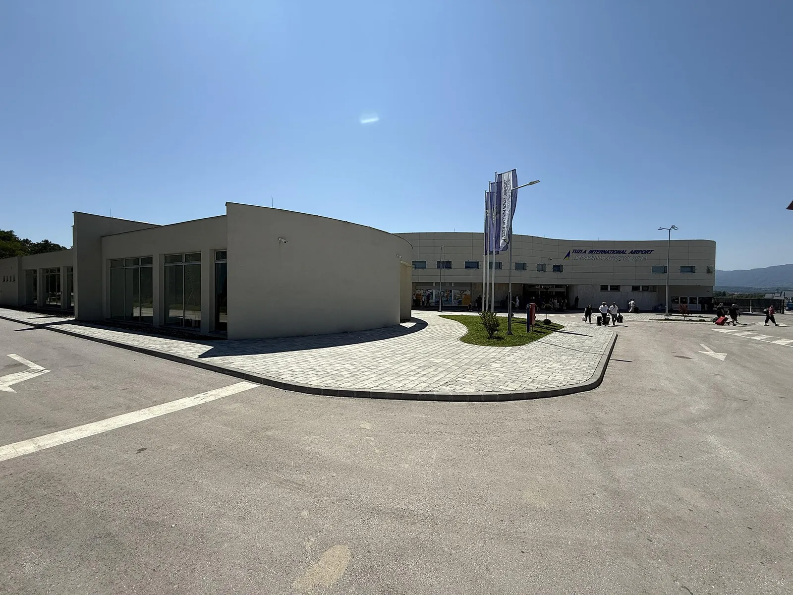 Low cream-coloured terminal building with a curved facade, flagpoles and a small lawn in front, passengers with suitcases walking across the forecourt under a clear blue sky