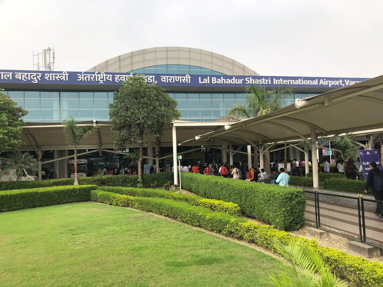 Landside kerb of Lal Bahadur Shastri International Airport, Varanasi, with the bilingual airport name on the glass terminal.