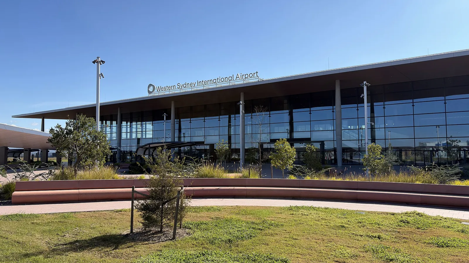 Long glass terminal facade of Western Sydney International Airport with the airport name on the roof edge, seen across a lawn under a clear sky