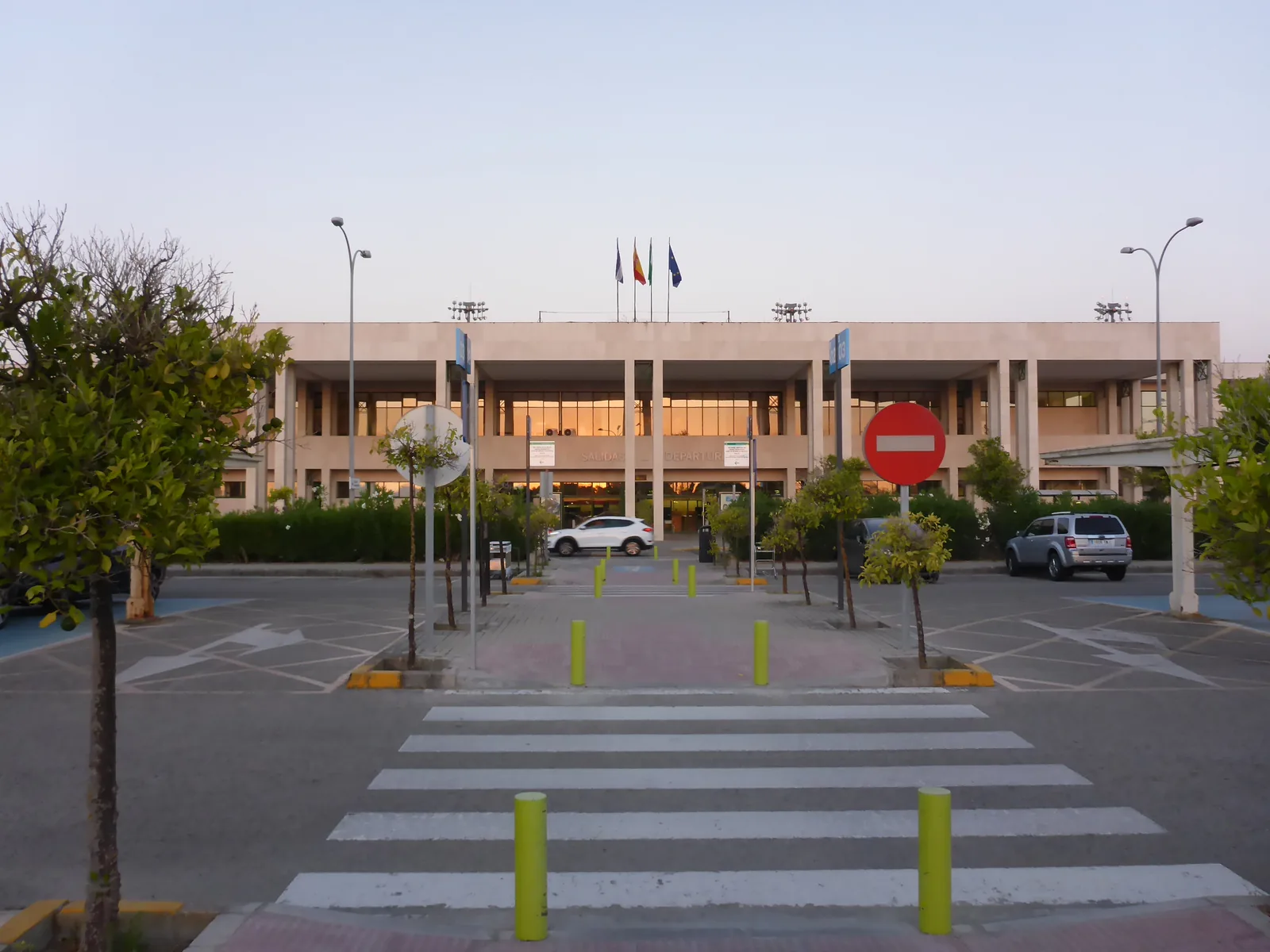 Front of the two-storey passenger terminal at Jerez Airport seen across the zebra crossing from the car park, with flags and the Aena sign