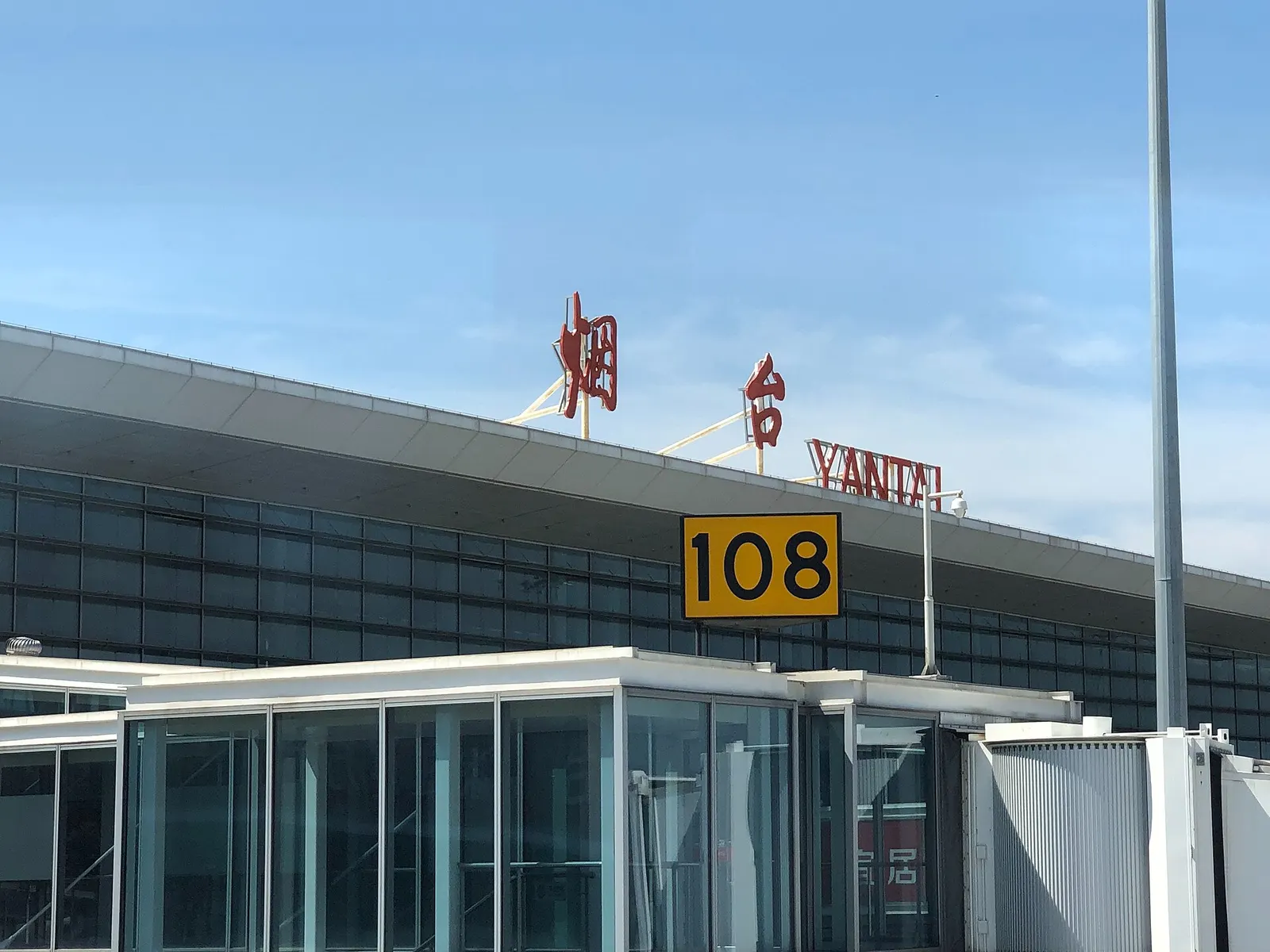 Closed 2015 T1 landside roof at Yantai Penglai on 25 Jun 2019: red 烟台 and YANTAI letters over a glass curtain wall, with yellow stand 108 on a jet bridge. Live passengers use T2 from 28 Jun 2024.