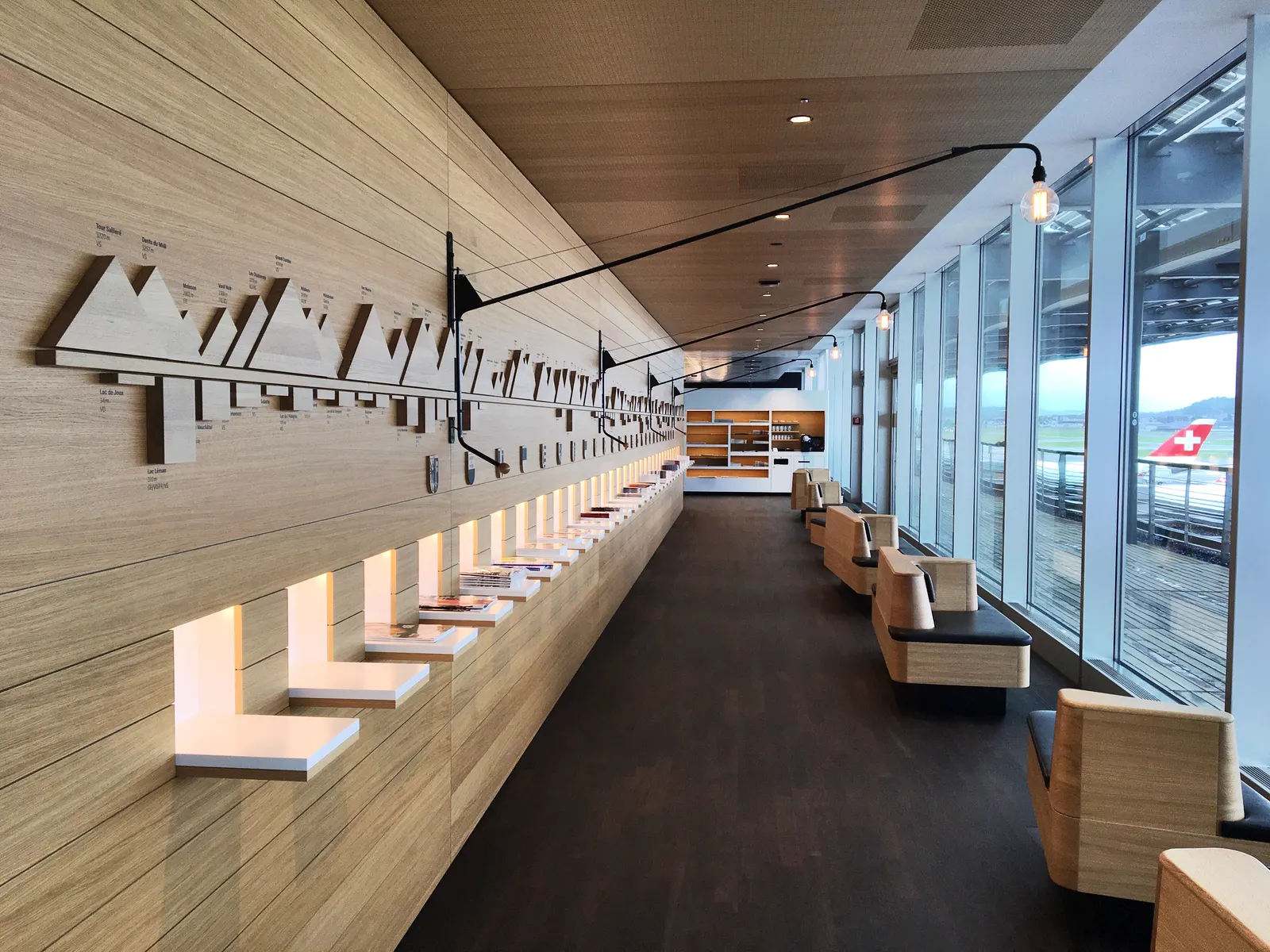 Wood-clad corridor with Alpine mountain relief and runway-facing seating inside a SWISS lounge at Zurich Airport (ZRH).